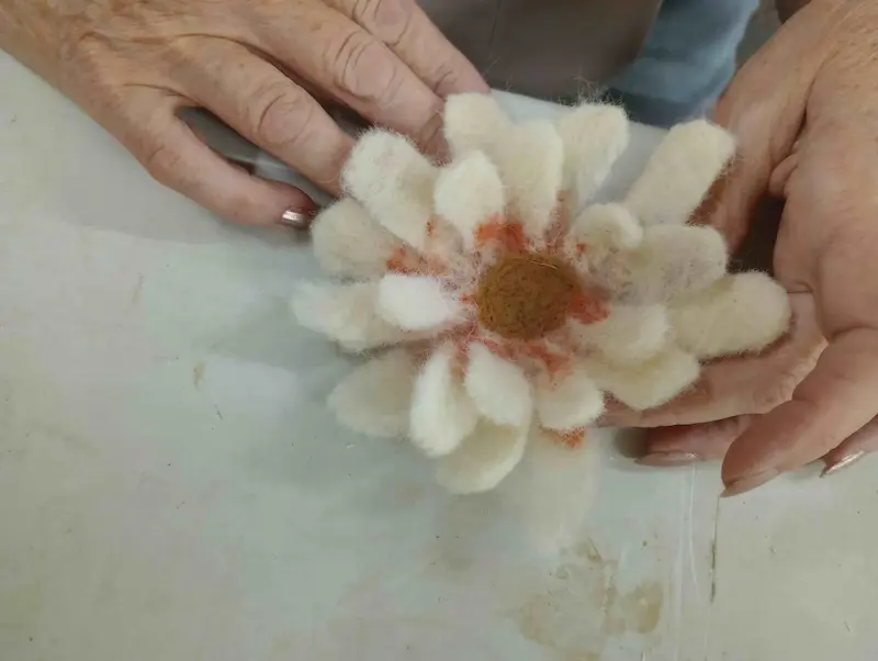 A pair of hands holding a handmade felt flower with white petals, orange accents, and a brown center, against a light-colored background.