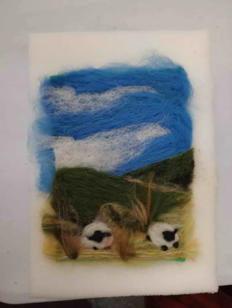 A felt artwork showing a landscape with a blue sky, white clouds, green hills, and two black and white sheep grazing on grass, made from colored wool fibers on a white background.