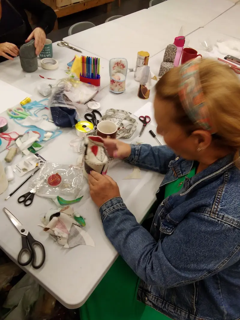 A group of people sitting around a table engaged in a craft activity, with materials like scissors, fabric, glue, and decorations scattered across the surface. One person is working on a decorated cylindrical object.
