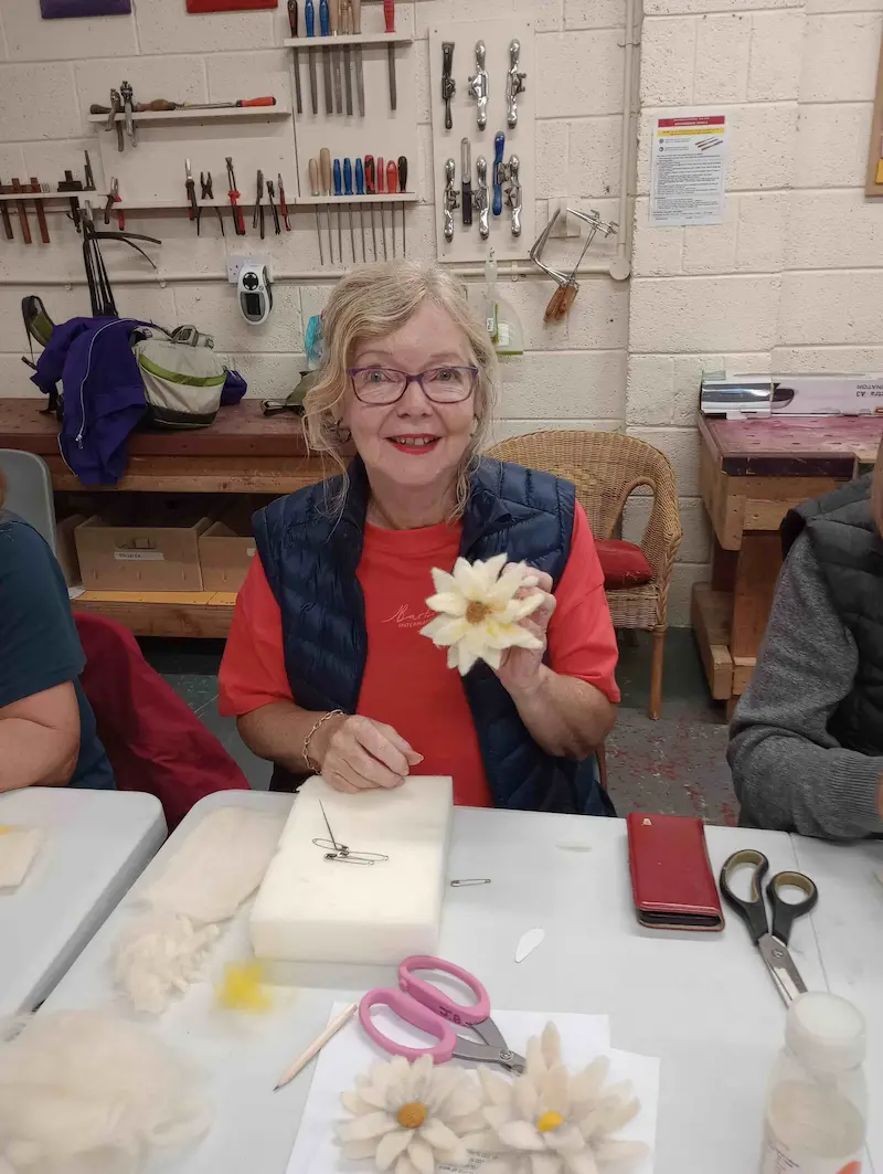 A person sitting at a table in a craft room, holding a handmade flower. The table is covered with crafting materials like scissors, felt pieces, and tools. The background shows shelves and a wall with hanging tools.