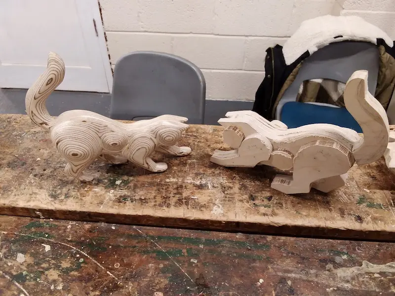 Two wooden cat sculptures made from layered wood pieces with a rough, unfinished look, placed on a workbench.