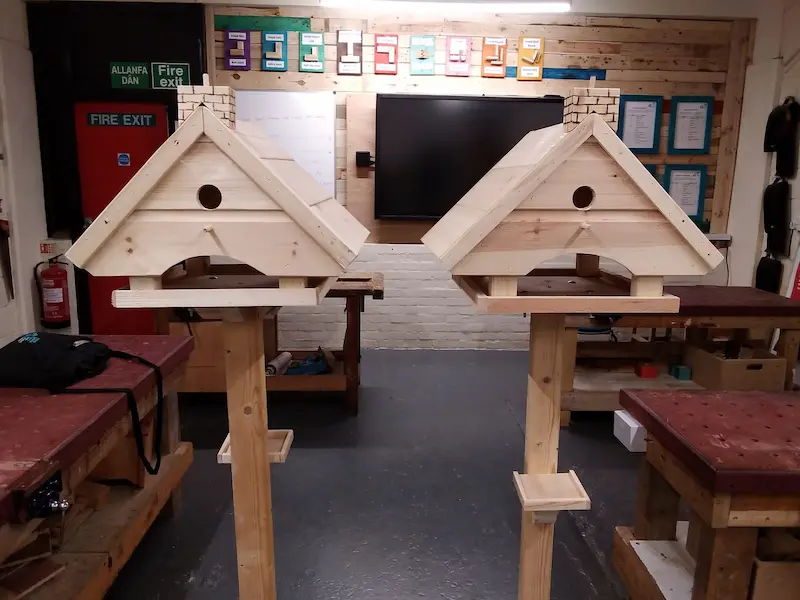 Two wooden birdhouses mounted on wooden stands in a workshop or classroom, with a red fire exit door, a blackboard or screen, and colorful posters and papers on the wall in the background.