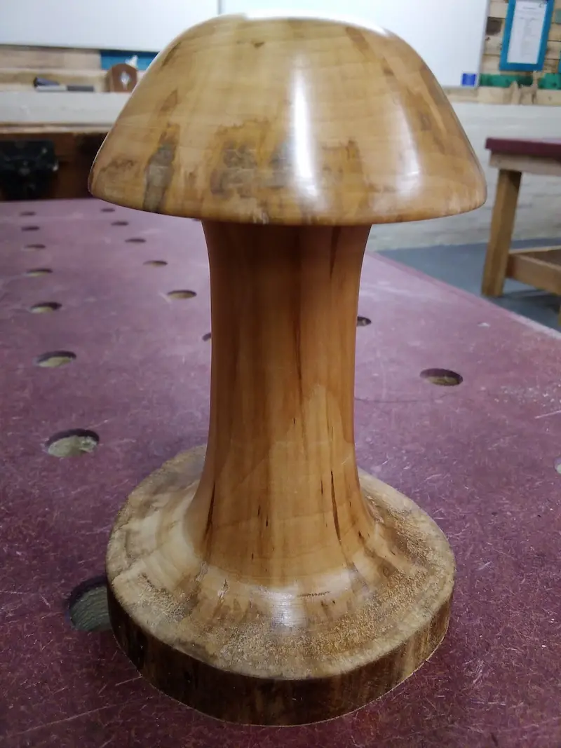 A wooden object shaped like a mushroom, with a wide, rounded top and a flared stem, placed on a perforated workbench in a woodworking shop.