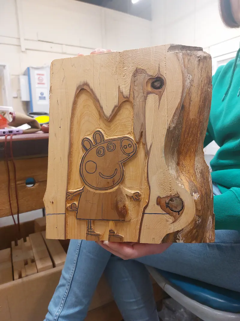 A wooden block featuring an engraved drawing of Peppa Pig, held by a person wearing blue jeans and a green shirt.